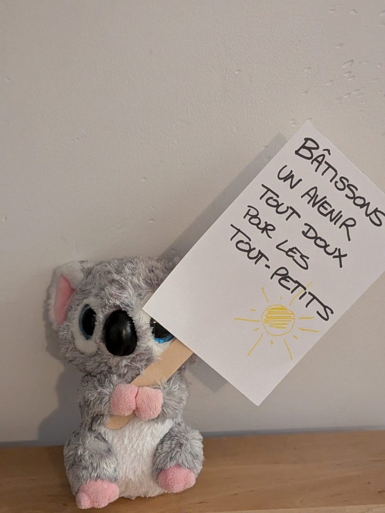 Stuffed koala holding up a sign reading “Bâtissons un avenir tout doux pour les tout-petits” (Let’s build a gentle future for our little ones)